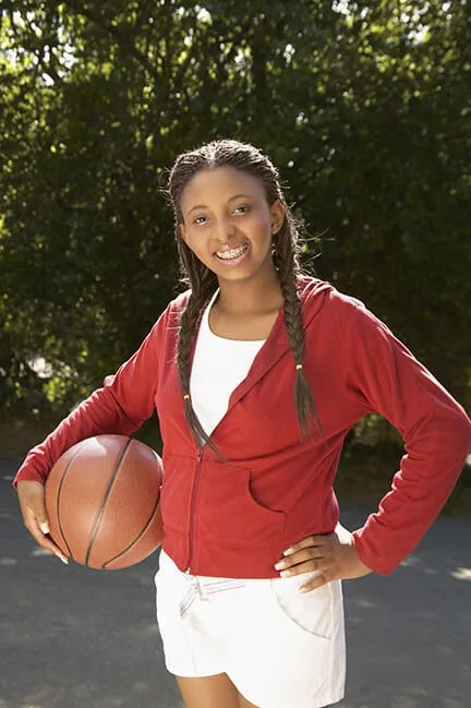 A young athlete in a red sweatshirt poses for a photo with her basketball, her braces clearly visible in her smile.